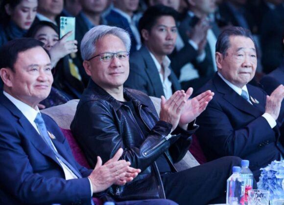 Three men in suits sitting and clapping at a formal event with a crowded audience in the background