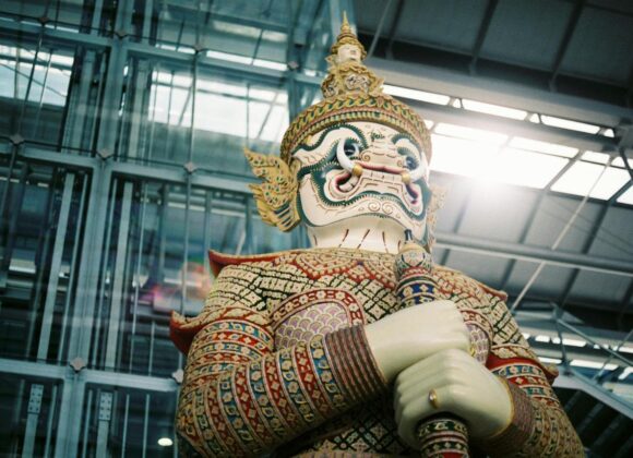 Colorful guardian demon statue indoors, hands folded around a staff, with an ornate headdress and patterned armor.