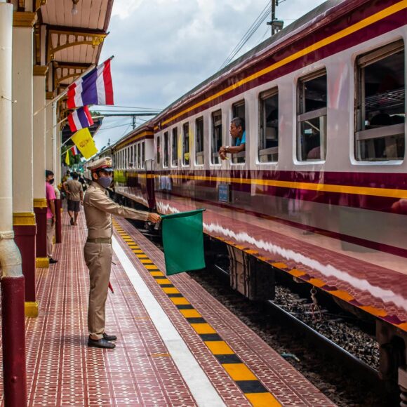 Train conductor in uniform signals a train with a green flag at a colorful station platform, passengers nearby and a maroon-yellow train alongside the tracks.