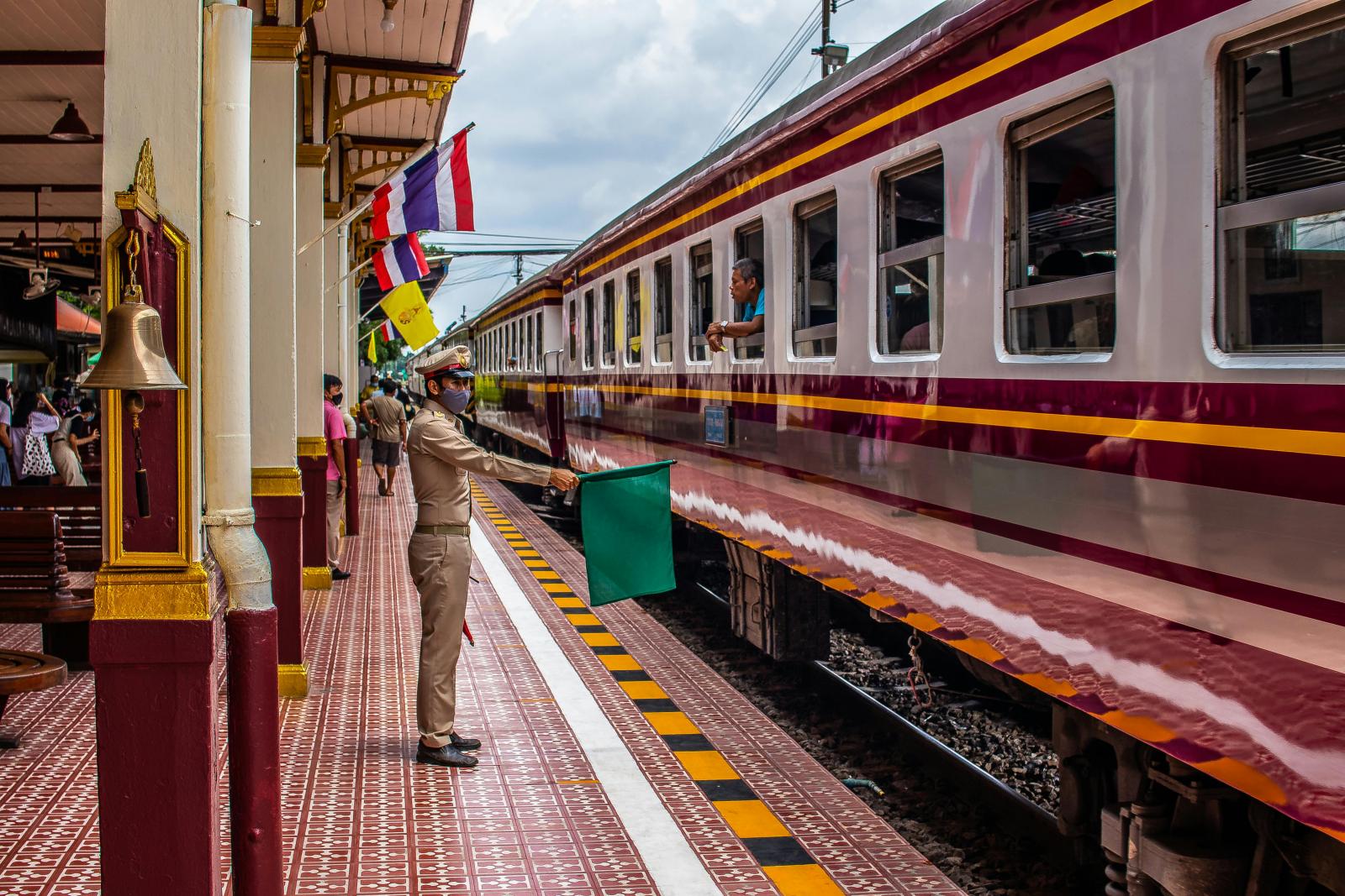 Bangkoktraincrashbusmakkasancrossing 00 可愛曼谷 narak Club Train Conductor in Uniform Signals a Train with a Green Flag at a Colorful Station Platform Passengers Nearby and a Maroon yellow Train Alongside the Tracks